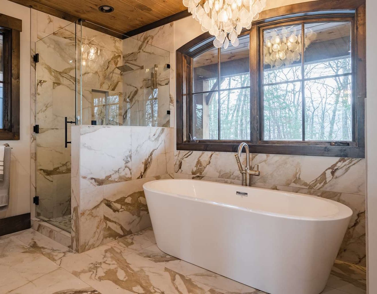 Luxury bathroom with marble surfaces, glass shower, freestanding tub, and forest view through a large window.